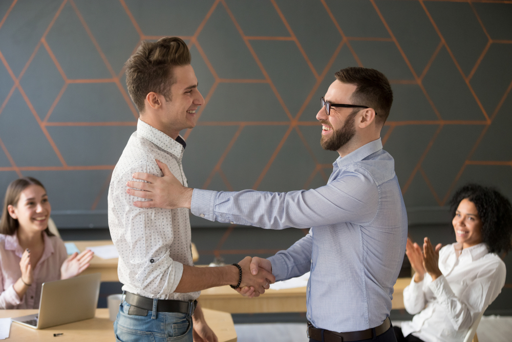 male employer shaking hands with male employee in front of the team